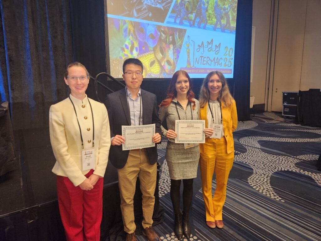 Four individuals in front a presentation screen. The two persons in the middle holding up their award certificate. 